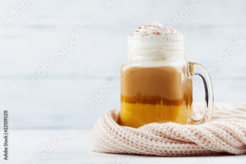 Pumpkin spiced latte or coffee in glass jar decorated knitted scarf on white wooden table. Autumn, fall or winter hot drink.