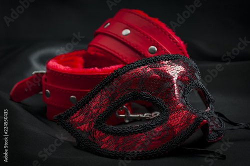 BDSM, bondage play, fetish wear and kinky sex toy concept with close up on erotic mask and red handcuffs isolated on black silk background