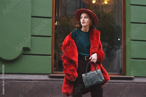  Outdoor fashion portrait of young beautiful confident lady wearing trendy orange faux fur coat, hat, green sweater, holding stylish snakeskin textured handbag, posing in street of city. Copy space