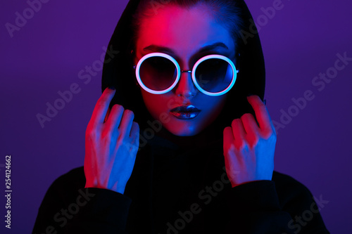 Portrait of fashion young girl in a black sweater with a hood and round sunglasses in red and blue neon light in the studio