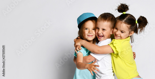 Studio portrait of children on a light background: full body shot of three children in bright clothes, two girls and one boy. Triplets, brother and sisters. hugging on camera. Family ties, friendship