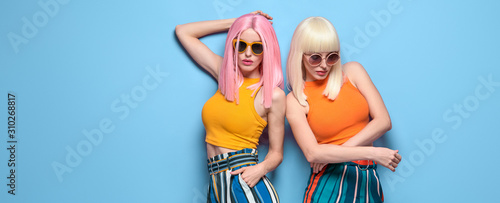 Two Young carefree Woman in stylish sunglasses, striped pants. Beautiful fashionable model girl in trendy summer outfit. Graceful friends with fashion dyed hairstyle, make up on blue. Creative banner