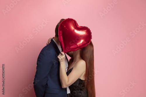 Happy holding balloons shaped hearts. Valentine's day celebration, happy caucasian couple on coral background. Concept of human emotions, facial expression, love, relations, romantic holidays.