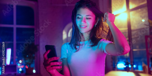 Taking selfie. Cinematic portrait of handsome stylish woman in neon lighted interior. Toned like cinema effects in purple-blue. Caucasian model using smartphone in colorful lights indoors. Flyer.