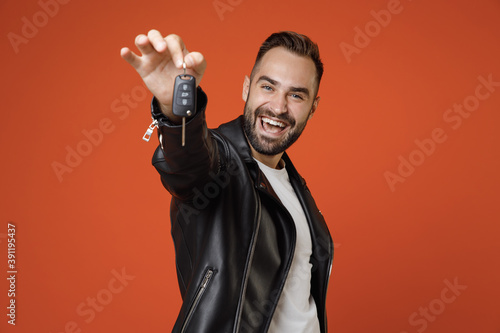 Cheerful successful young bearded man 20s wearing basic white t-shirt, black leather jacket standing hold in hands car keys looking camera isolated on bright orange colour background studio portrait.
