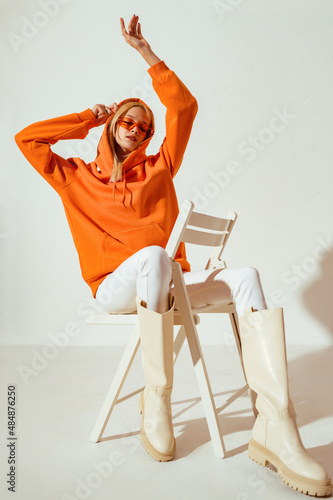 Full-length studio fashion portrait of playful confident woman wearing stylish orange hoodie, sunglasses, white skinny jeans, high boots, sitting, posing on chair. 