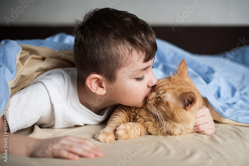 The boy hugs a red cat on a bed under a blanket