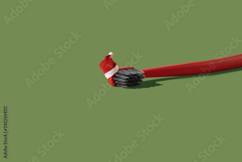 Single red toothbrush with red and white Santa hat laying on green background with copy space and shadow. Dental medical Christmas and New Year concept. Creative health still life levitation.