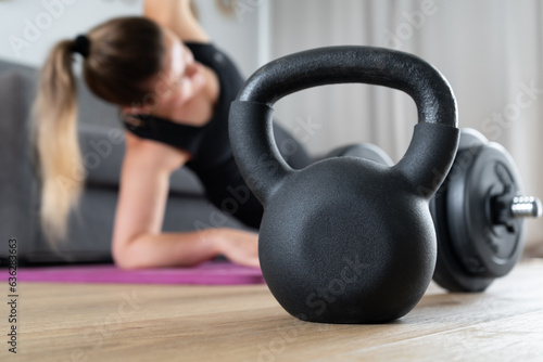 Woman exercising at home in the living room on yoga mat. House fitness workout, sport training concept. Focus on gym exercise equipment, kettlebell, dumbbell barbell. Female doing pilates, stretching.