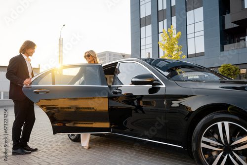 Business man helps business lady to get in the car, opening vehicle door near office building on sunset. Concept of business lifestyle and transportation