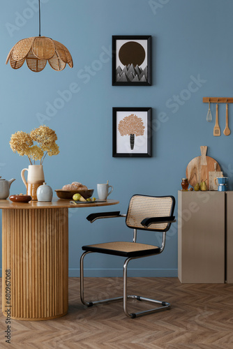 Warm and cozy dining room and kitchen interior with mock up poster frame, round table, rattan chair, beige commode, blue wall, vase with flowers and kitchen accessories. Home decor. Template.