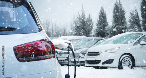 Close up of electric car with a connected charging cable on the blurred background of a snow covered parked cars.