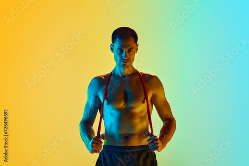 Handsome young man with muscular, shirtless, relief body posing wit fitness resistance band against gradient blue yellow background in neon light. Concept of active and healthy lifestyle, sport