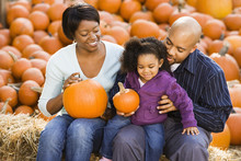 Happy Family Sitting On Hay Bales And Holding Pumpkins.