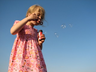 little girl blowing away soap bubble against a clear blue sky.