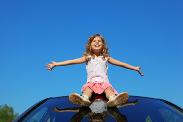 little girl sitting on car roof with open hands on blue sky