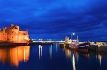 Fishing Boat In The Harbor At Night Beside Illuminated Castle