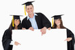 © WavebreakmediaMicro - Three graduates pointing to the blank sign as they look at the c