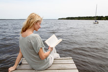 Woman Reading Book Sitting On Lake Boardwalk