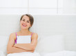 © Alliance - Happy young woman sitting on bed and embracing tablet PC