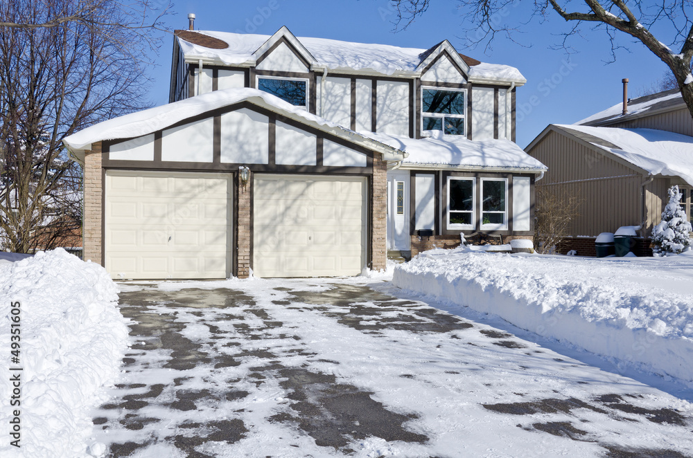 A two-story suburban house with a double garage and a neatly cleared driveway surrounded by deep snow.