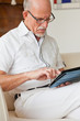 © ysbrandcosijn - Senior man with glasses using tablet on couch in living room.