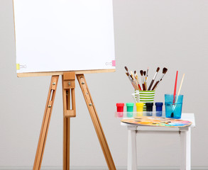 Wooden easel with clean paper and art supplies in room