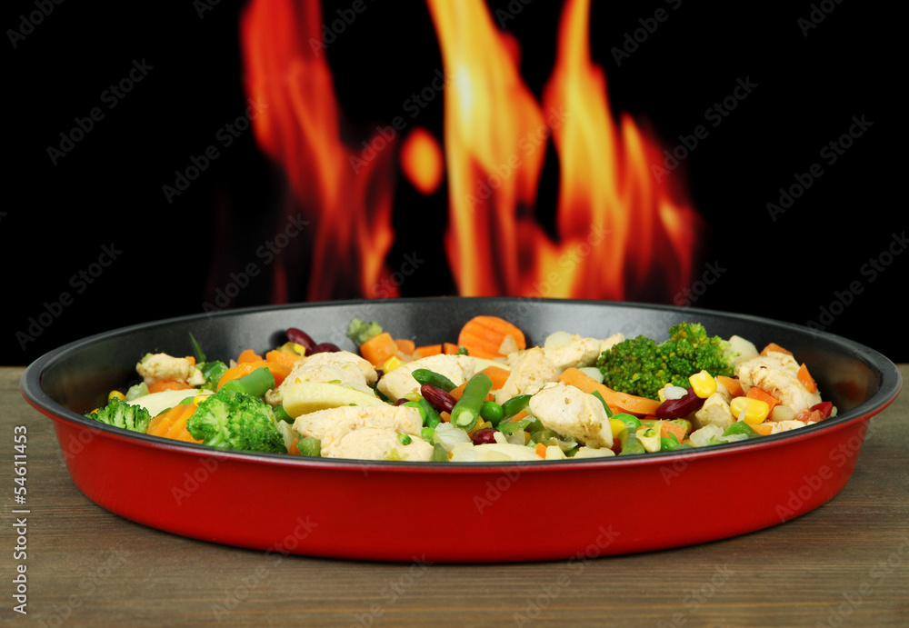 Casserole with vegetables and meat on pan, on fire background