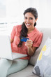 © lightwavemedia - Excited brunette sitting on her sofa using laptop to shop online