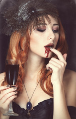 Redhead vampire woman with glass of blood. Photo in vintage styl