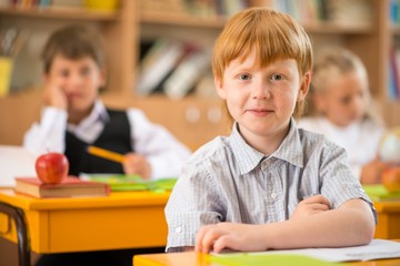 Fototapete - Little redhead schoolboy behind school desk