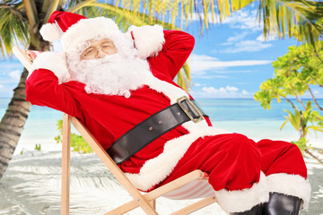 Wall Mural - Relaxed Santa Claus seated on a chair, on a beach with palm tree