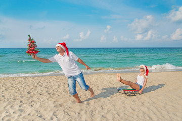 Sticker - Young couple running at sea beach in santa hats with sled and ch