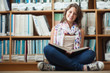 © WavebreakMediaMicro - Smiling female student against bookshelf reading a book on the l