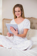 © lightwavemedia - Natural smiling woman sitting on bed using tablet