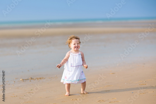 Baby Girl With Curly Hair In A White Dress Running On A Beach