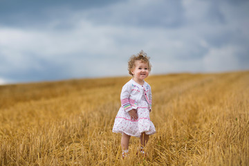 Wall Mural - Curly baby girl walking in a golden wheat field at sunset