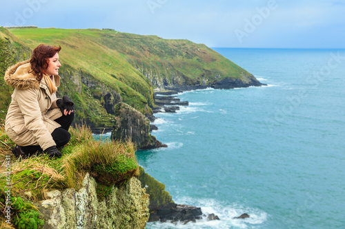 Fotografie, Obraz Woman sitting on rock cliff looking to ocean Co. Cork Ireland