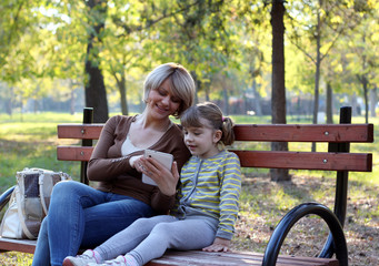 Wall Mural - mother and daughter sitting on bench and play with tablet