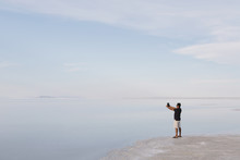 A Man Standing At Edge Of The Flooded Bonneville Salt Flats At Dusk, Taking A Photograph With A Tablet Device, Near Wendover.