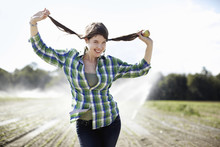 A Girl In A Green Checked Shirt With Braids Standing In A Field With Sprinklers Working In The Background.