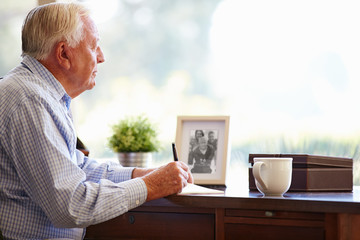 Senior Man Writing Memoirs In Book Sitting At Desk