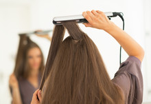 Young Woman Using Hair Straightener In Bathroom. Rear View