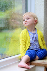 Wall Mural - Cute little girl looking through window on rainy day