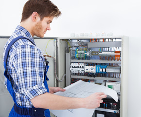 Technician Analyzing Blueprint In Front Of Fusebox