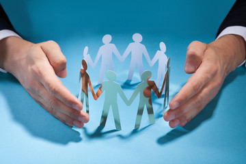 Businessman's Hands Protecting Team Of Paper People On Desk