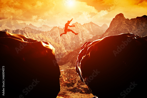 Láminas Man jumping over precipice between two mountains at sunset