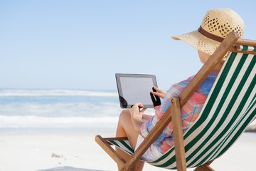 Woman in sunhat sitting on beach in deck chair using tablet pc