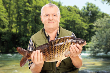 Wall Mural - Mature fisherman showing his catch standing by a river