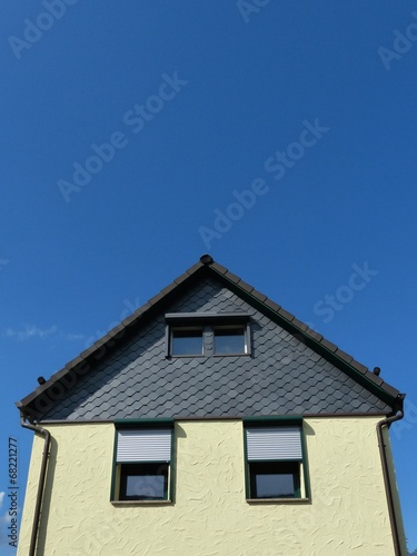 Fassade mit gelbem Putz und Schindeln aus grauem Schiefer vor blauem